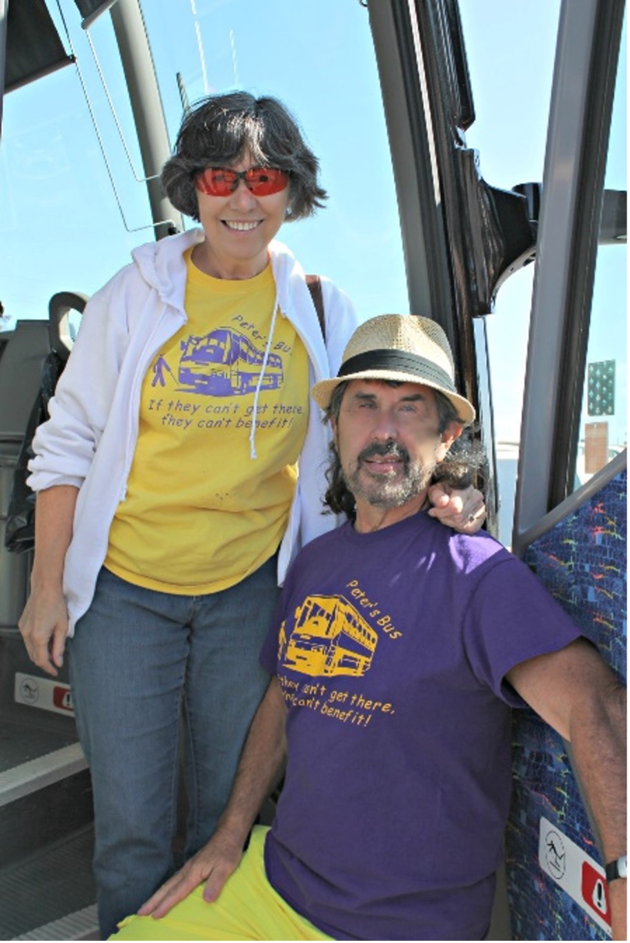 Peter and his wife Denise pose for a photograph at the bus entrance. Both are wearing t-shirts that say, "If they can't get there, they can't benefit."