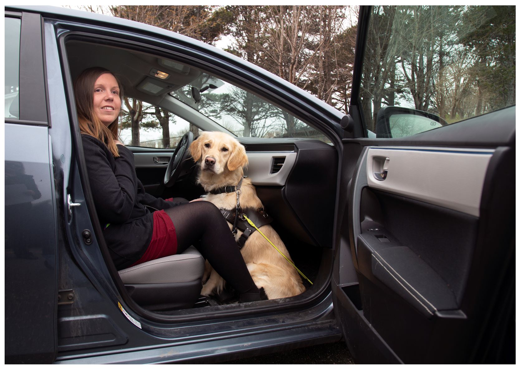 A woman is sitting in the front passenger seat of a car with the door open; her guide dog, a golden retriever, is sitting at her feet in the passenger footwell.