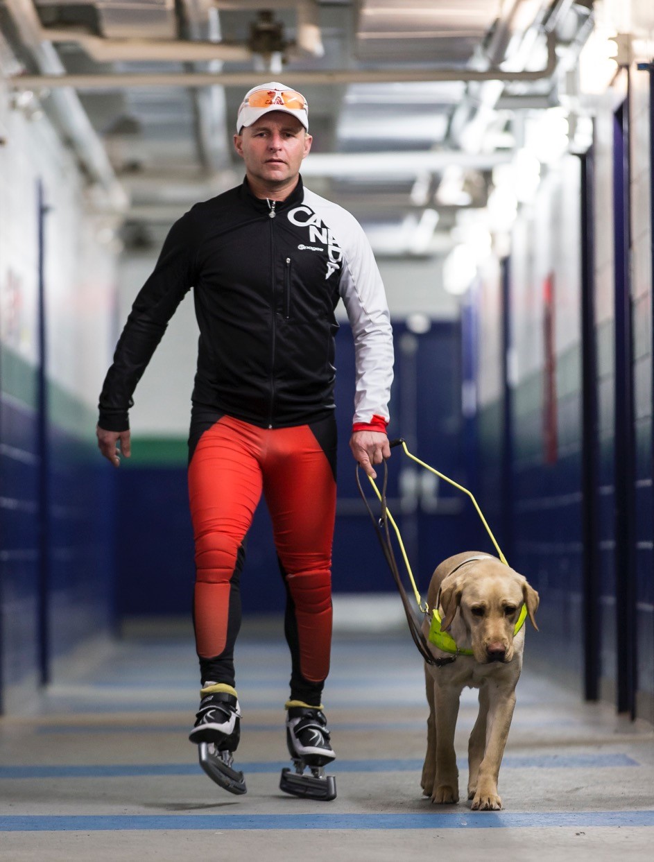 Kevin wearing ice skates and sports clothing walking down a corridor with his guide dog.