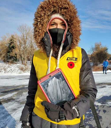Une collectrice de fonds portant une veste jaune d'INCA, tenant une tablette sécurisée et portant un masque facial, est posée debout, emmitouflée pour se protéger du temps froid