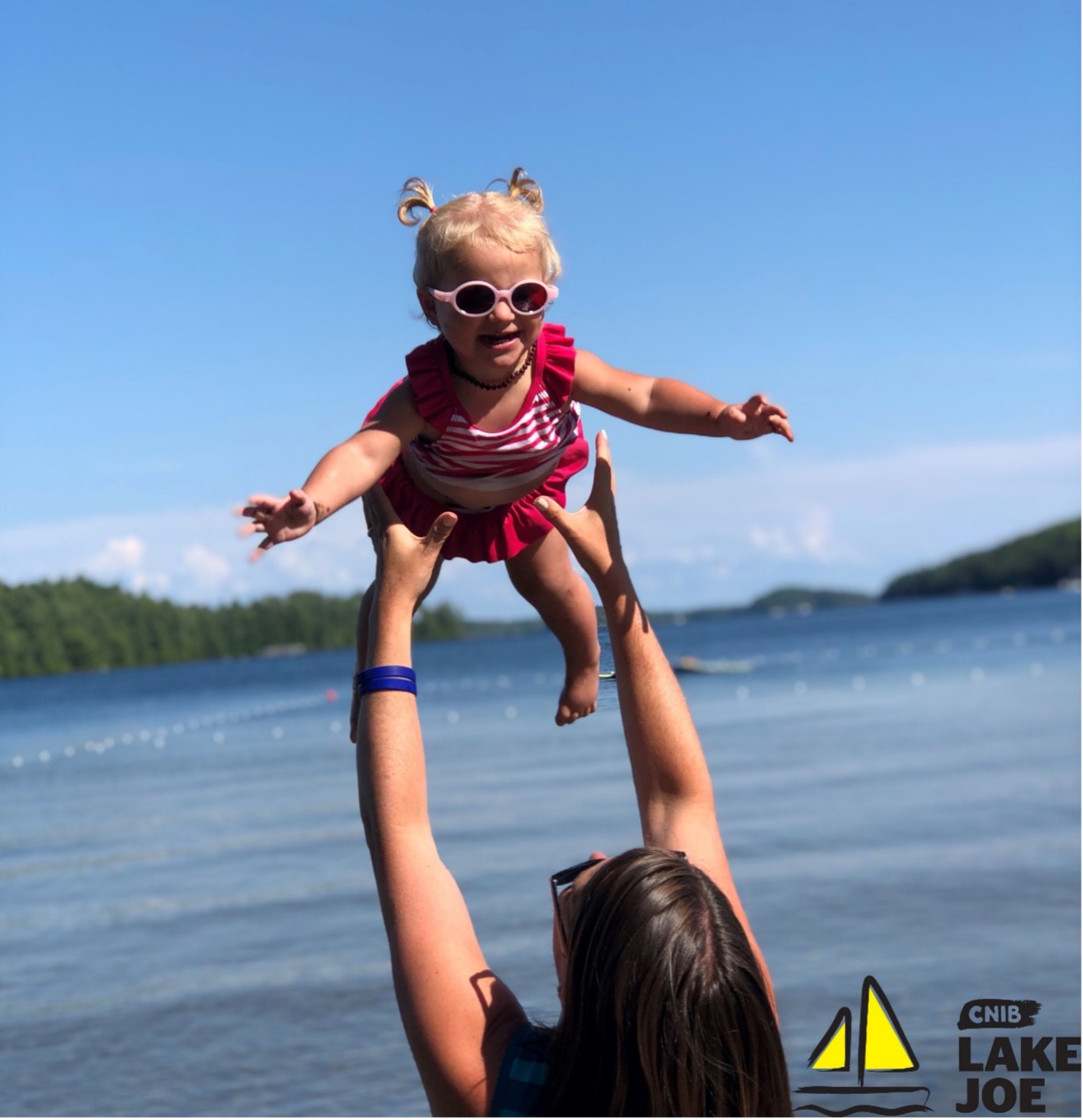 A little girl wearing a bathing suit and sunglasses is being thrown in the air by her mom on the shore at CNIB Lake Joe. CNIB Lake Joe yellow and black sailboat logo at the bottom-right corner of the image.