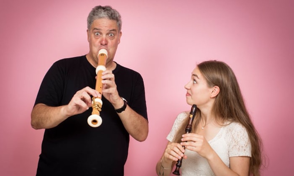 A man and a woman are playing recorders. The man has a funny look on his face, and the woman is smiling