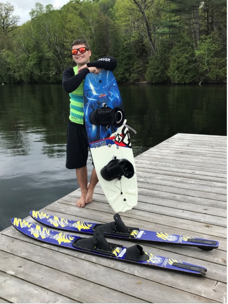 Josh is standing on the edge of the dock wearing a black shirt and shorts with a green life jacket. A set of blue and yellow water skiis on the dock, and he’s leaning against a wakeboard. He’s wearing sunglasses and smiling.