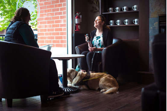 Larissa is sitting in a chair across from another person in a coffee shop by the window with her guide dog Piper laying on the floor at her feet.