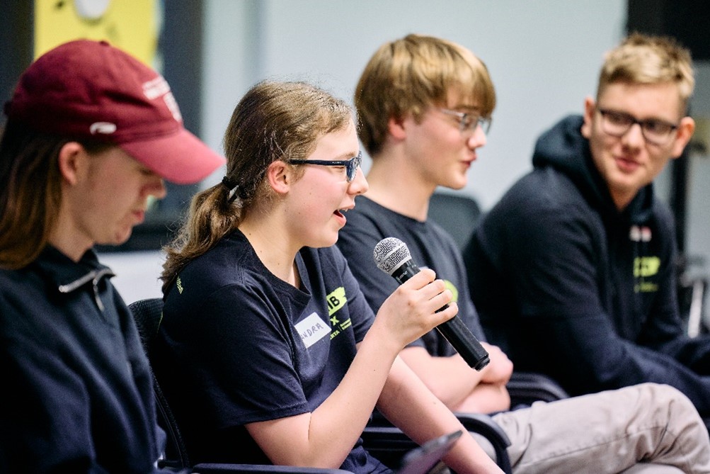 Three youths, each with light skin, sit in a row. The young girl on the left speaks into a microphone.