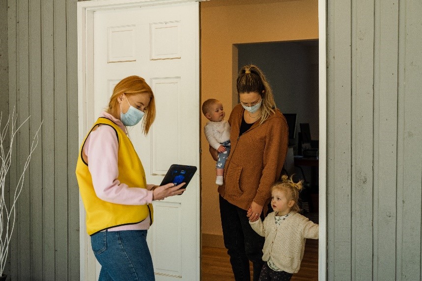 Une collectrice de fonds d'INCA, vêtue d'une veste et d'un masque jaune, cherchant dans sa tablette, debout devant la porte d'une donatrice qui tient ses deux jeunes enfants
