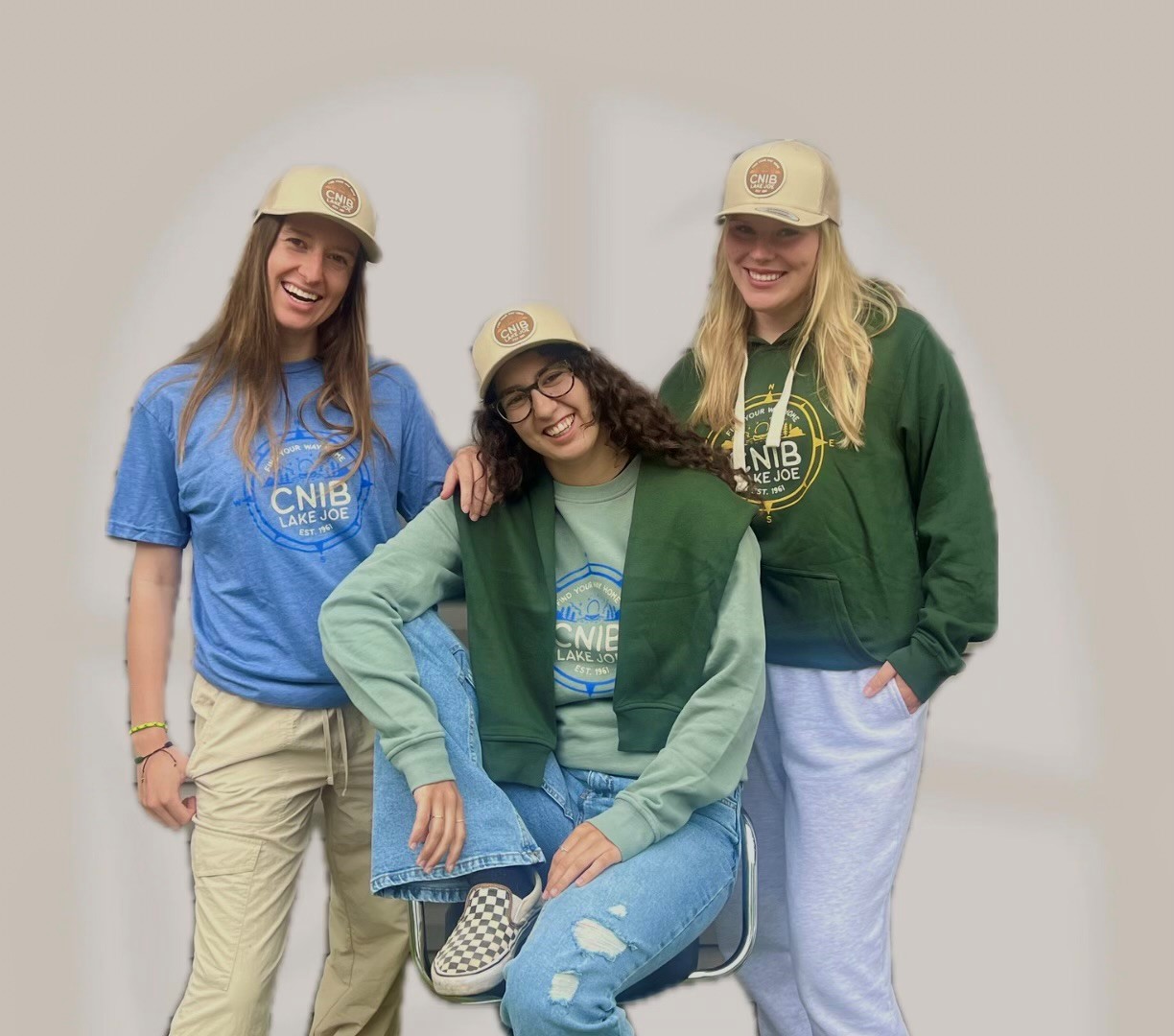 Three young women are posing wearing tan coloured baseball hats with CNIB Lake Joe logo on them, as well as different colour sweatshirts (light blue and mint green) and hoodies (dark green).