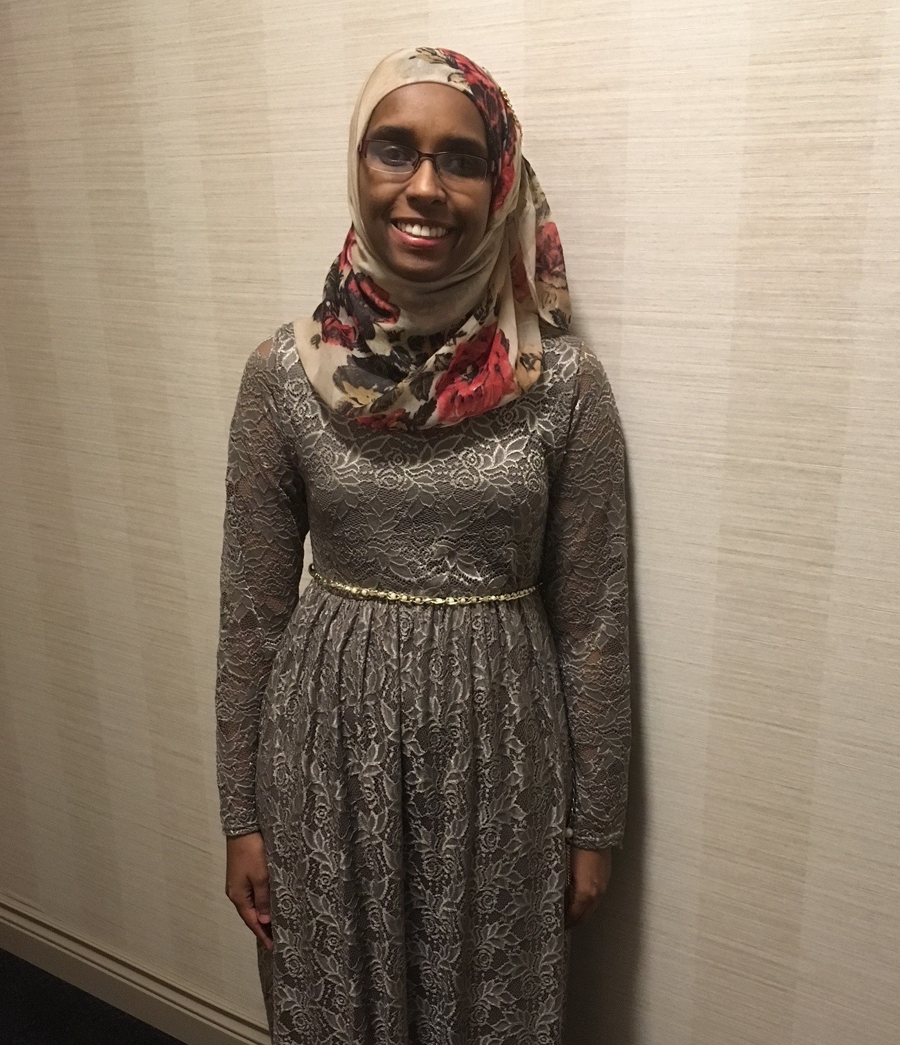 Ramla sourit et pose pour une photo sur un fond blanc. Elle porte une robe grise et un hijab.