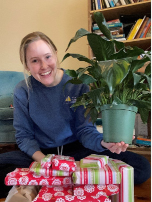 Amy est agenouillée devant une pile de cadeaux emballés et, tout sourire, tient une de ses plantes d'intérieur par-dessus les cadeaux.