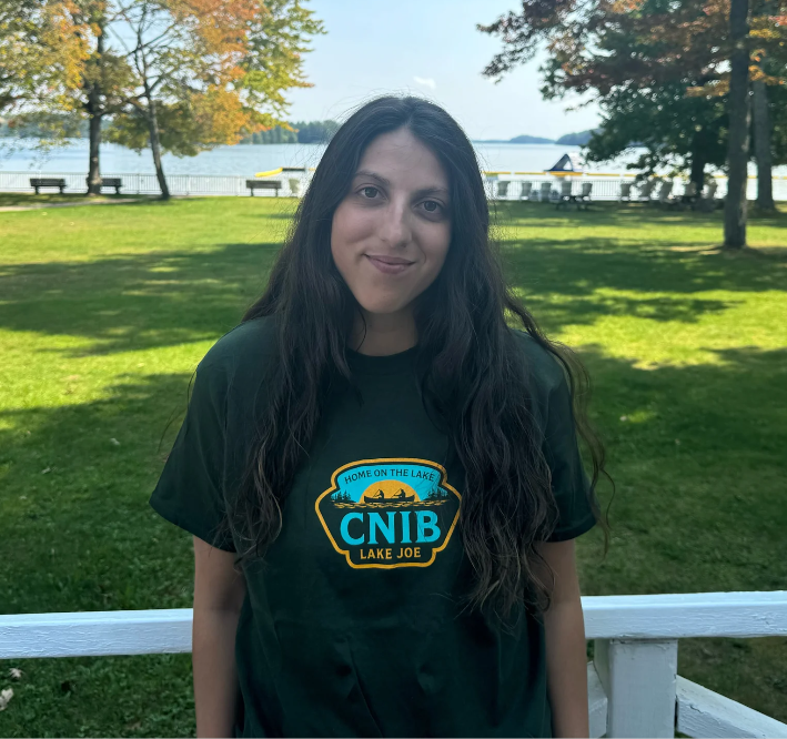 Stela is standing on the CNIB Lake Joe boardwalk with her back to the lake wearing a CNIB Lake Joe olive green t-shirt.