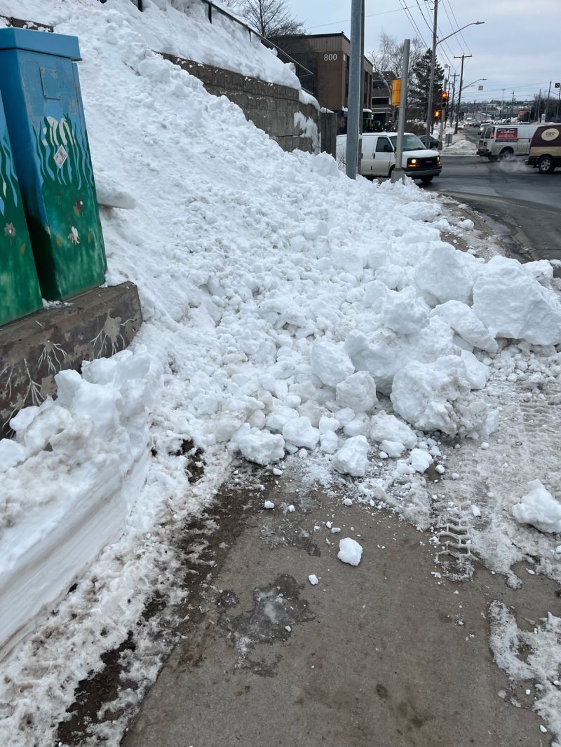 A snowbank blocking part of the sidewalk requiring pedestrians to step out on to road. A portion of cleared sidewalk that disappears under a large pile of snow roughly 3 feet on the left sloping to 2 feet on the right. 