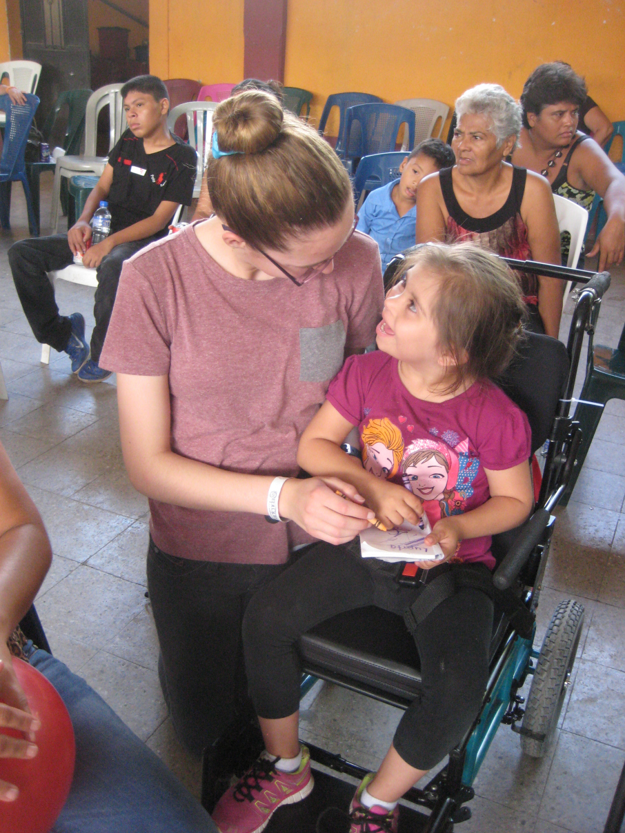 In Guatemala, a teenage Veronika crouches down next to a young girl in a wheelchair. They are colouring on a piece of paper. 