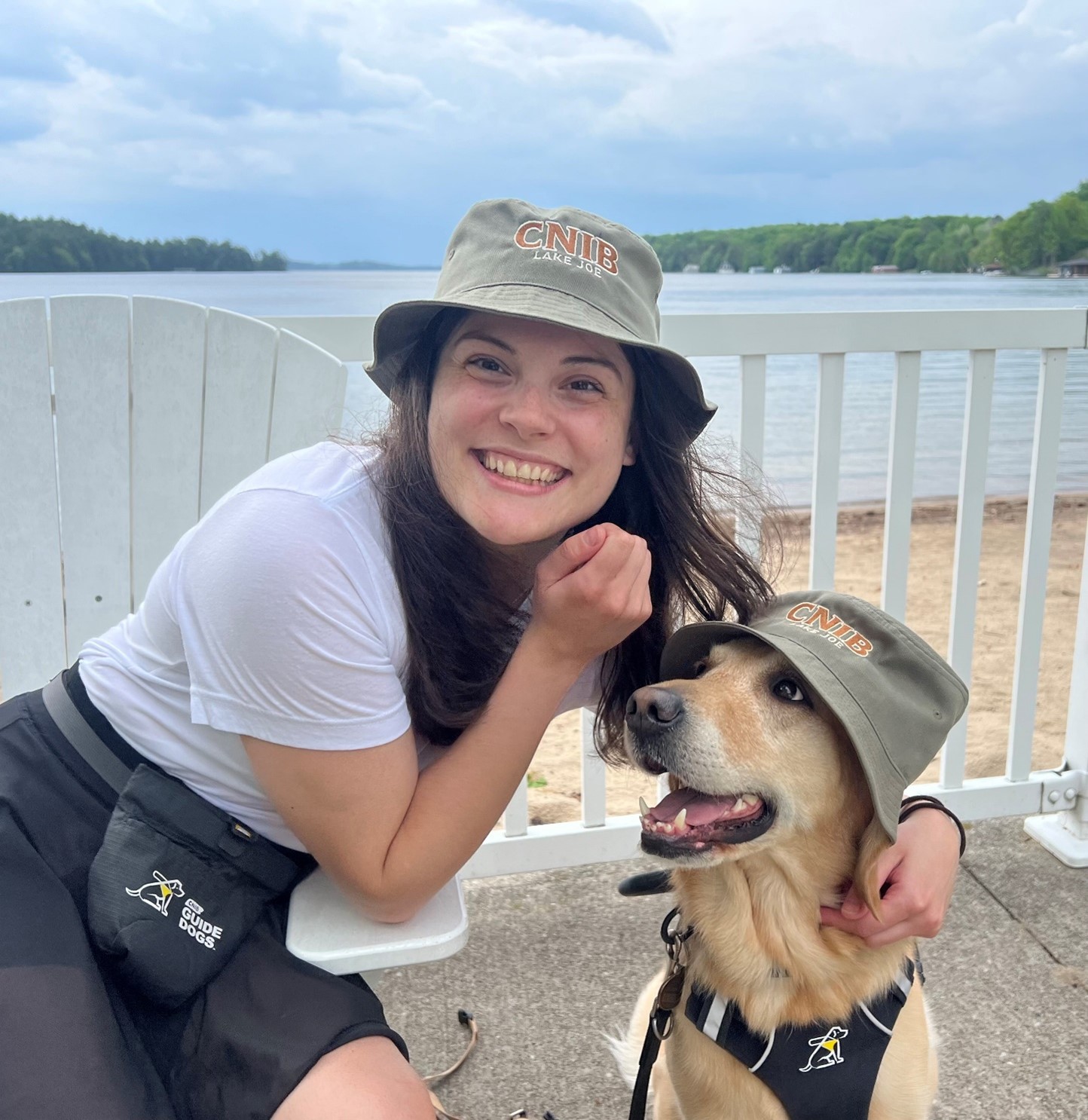 A young woman is sitting on a white Muskoka chair with Lake Joseph behind her. She has her arm around a yellow labrador retriever. Both are wearing CNIB lake Joe bucket hats. The woman is smiling at the camera and the dog is looking at the woman.