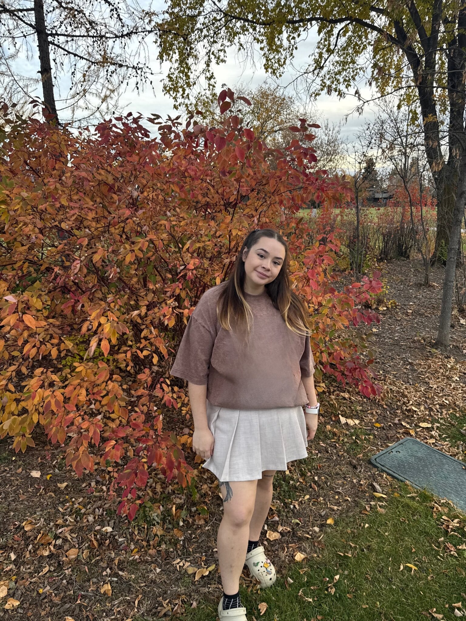 Alexis is standing in front of an autumnal bush wearing a brown t-shirt and beige skirt. She has long brown hair that fades into blond and she is standing with her head on a tilt smiling. 