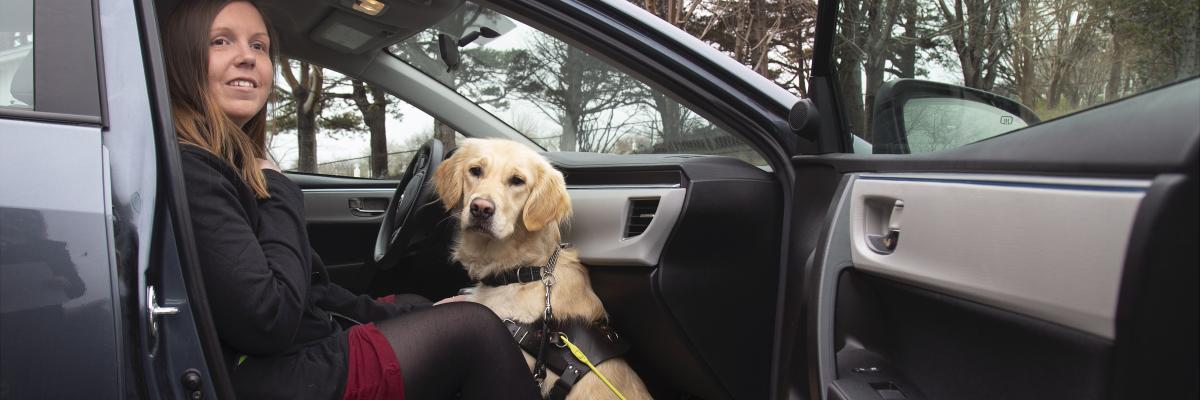 Kelly et de son chien-guide Maple, assis sur le siège côté passager d'une voiture la portière ouverte, souriant pour la caméra.