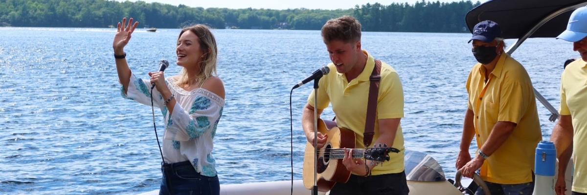 Leah Daniels chantant et saluant les spectateurs avec Will Hebbes à ses côtés jouant de la guitare sur un ponton scénique, divertissant les invités des quais sur le lac Muskoka. 