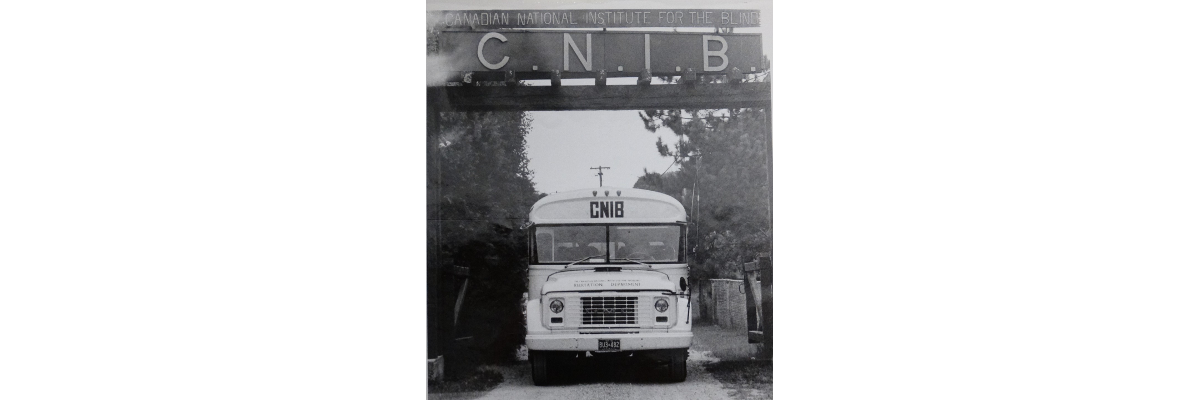 Une photo en noir et blanc d'un ancien autobus scolaire bien marqué CNIB  à l'avant, passant sous le panneau de bienvenue au Centre Lake Joseph d’INCA. 