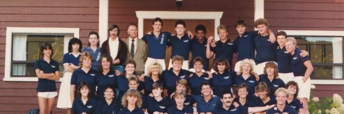 Photo de l'équipe du personnel de 1986 (35 hommes et femmes vêtus de chemises de golf bleues et de shorts blancs) posant à l'extérieur du bâtiment du personnel pour le 25e anniversaire du camp Lake Joe d'INCA. 