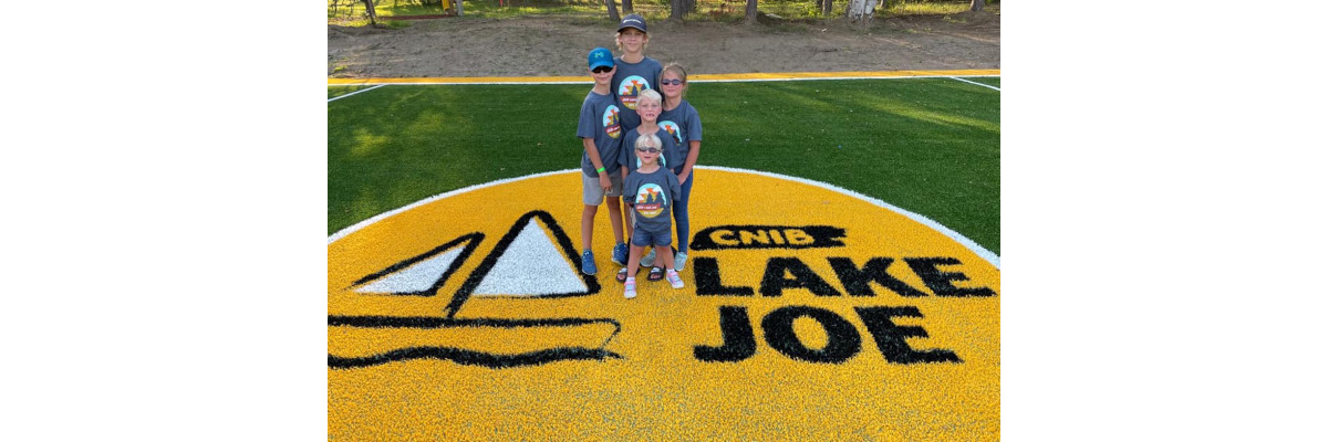 Cinq enfants posent sur le logo jaune géant d’INCA au milieu du nouveau terrain de soccer du camp Lake Joe. Ils portent tous un t-shirt gris à l’effigie du camp Lake Joe d’INCA