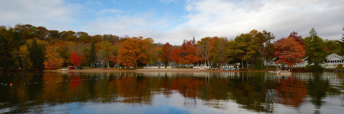 A beautiful autumn view of CNIB Lake Joe’s shoreline from the water. The foliage is changing colours on the trees lining the lake.