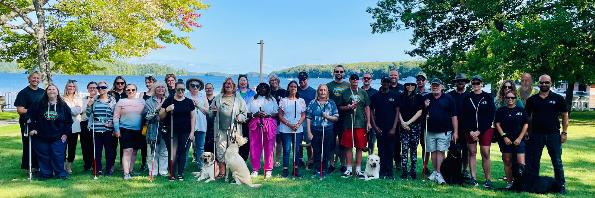 A group photo taken by the waterfront at CNIB Lake Joe. The group consists of members of the Vision Loss Rehabilitation team, the CNIB Guide Dogs team and participants of the Orientation and Mobility Immersion Program. 