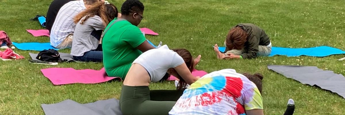 Eight people are seated on the grass on colourful yoga mats. They are all bent over with one leg stretched out. A woman is in the same pose across from them.