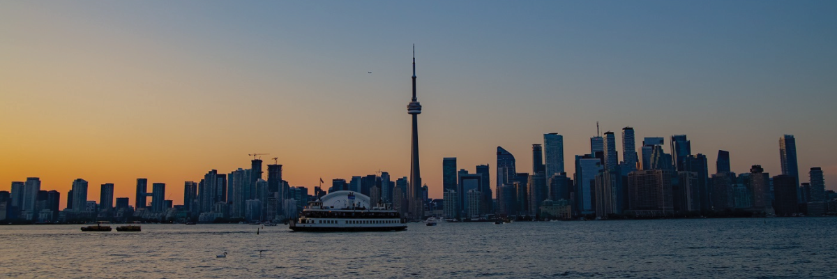 La ligne d'horizon de Toronto au coucher du soleil, prise depuis l'île de Toronto. Le ciel est bleu foncé, et la lumière du soleil, jaune et or, encadre les édifices et la ligne d'horizon. Un traversier avance dans le Port.