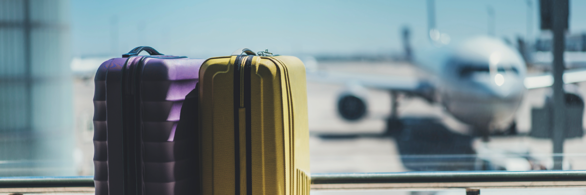 Two suitcases sit side-by-side in front of a window in an airport departure lounge. Outside the window, an airplane sits on the tarmac at the boarding gate. The airplane is out of focus and blurry.    