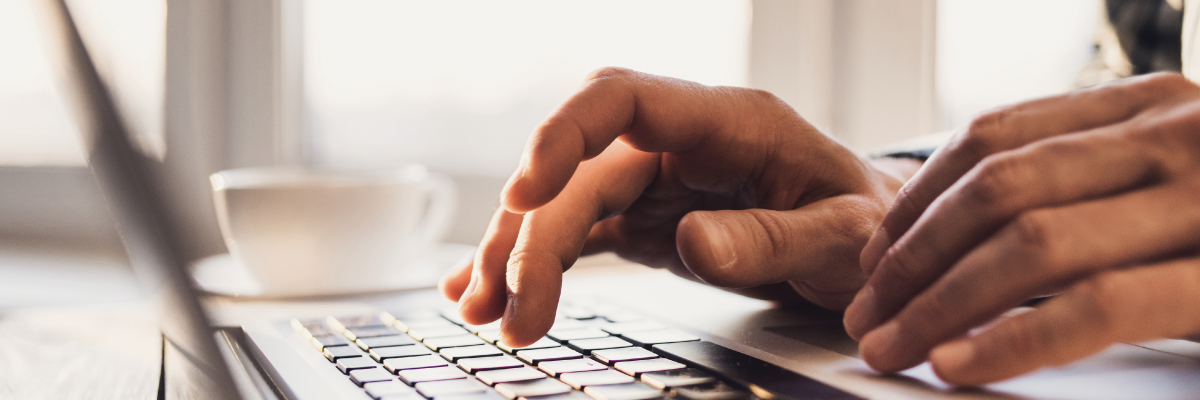 A close-up of a person’s hands typing on a laptop keyboard. The primary focus is on the hands and the keyboard, the background is blurred but it’s a well-lit indoor setting.