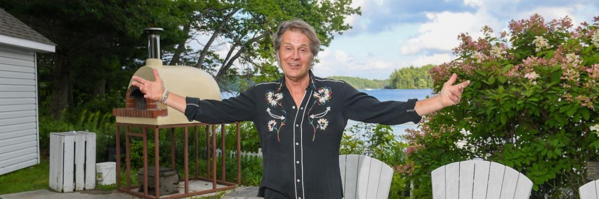 Jim Cuddy is standing outside in front of the pizza oven at CNIB Lake Joe. There are white Muskoka chairs behind him as well as bushes and Lake Joseph. There is a blue sky. Jim is wearing black jeans and a black cowboy shirt with white embroidery. He is standing with his arms outstretched and smiling.