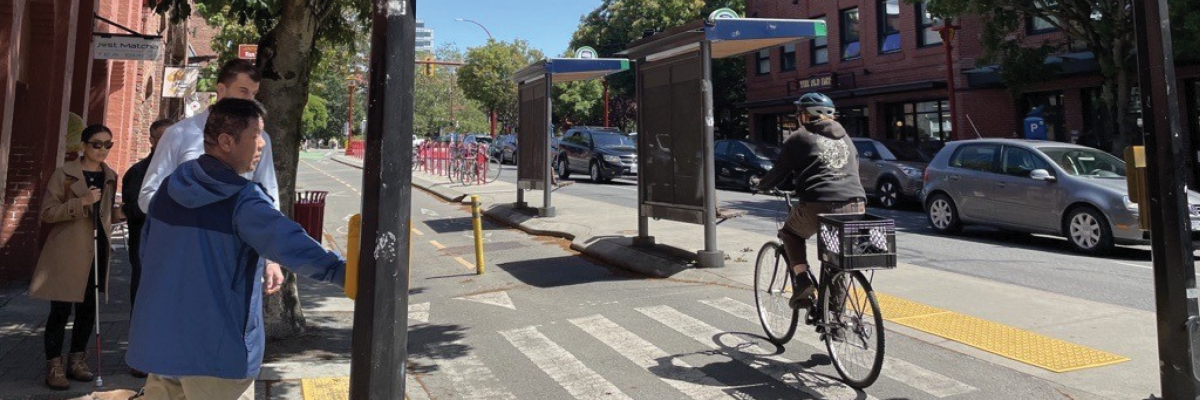 A pedestrian and his guide dog attempt to cross the street to a floating bus stop. The pedestrian presses a crosswalk button as a cyclist ride their bikes through the crosswalk.  