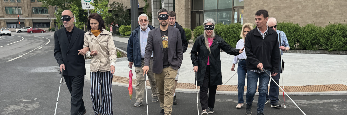 CNIB staff provided sighted guide to blindfolded city councillors. The group crosses the road. The councillors are carrying white canes to feel the texture of the road.