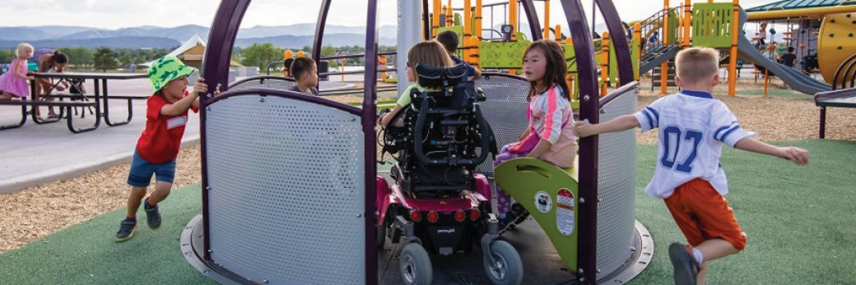 Children play on an accessible playground on a sunny summer day. A child who uses a wheelchair is on a wheelchair-accessible carousel with three other children. Two more children run and pull the carousel to make it spin for the children sitting on it.