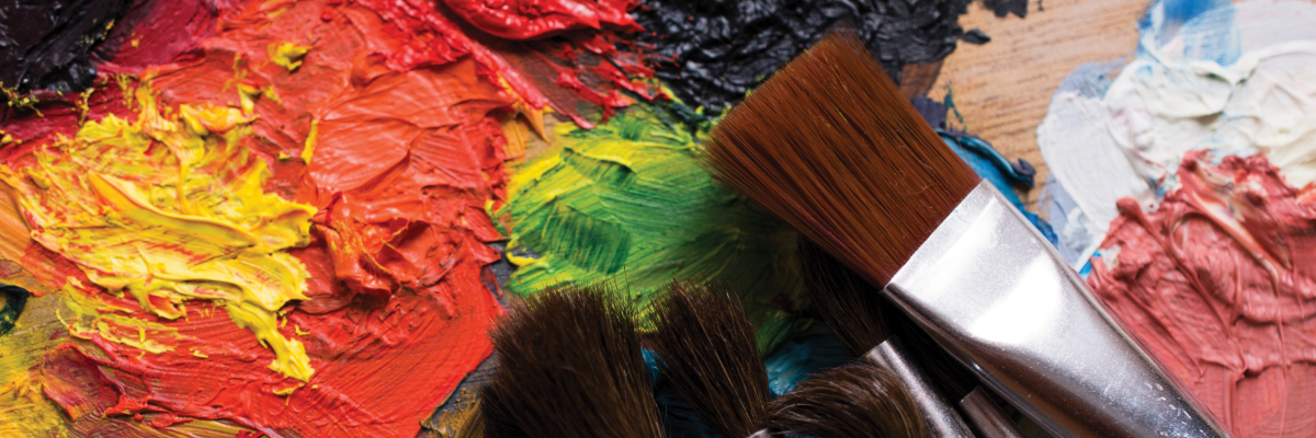 A colourful hand-painted mural with various paintbrushes lying on top of the mural. The abstract mural has thick, textured paint in vibrant colours like red, yellow and green.