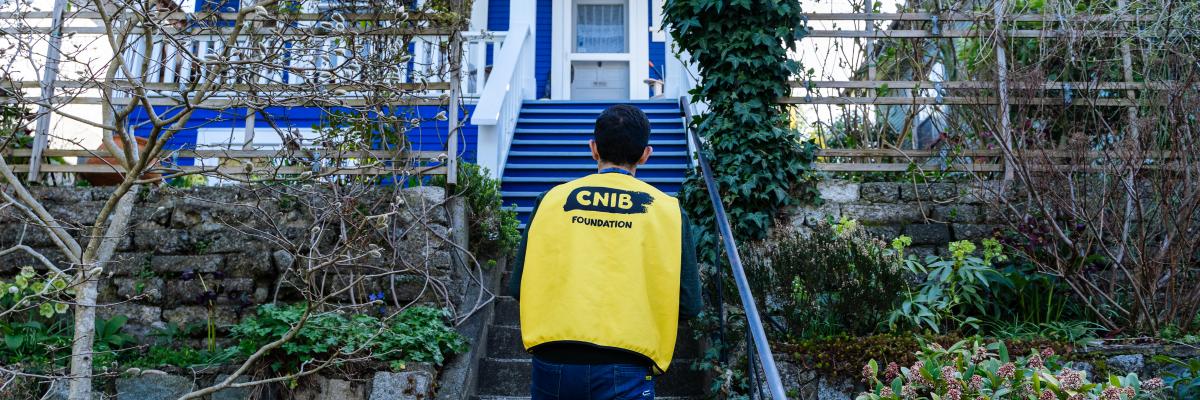 A canvasser wearing a yellow vest with the CNIB Foundation logo on the back ascends a flight of stairs, surrounded by a green garden, towards the front door of a house.