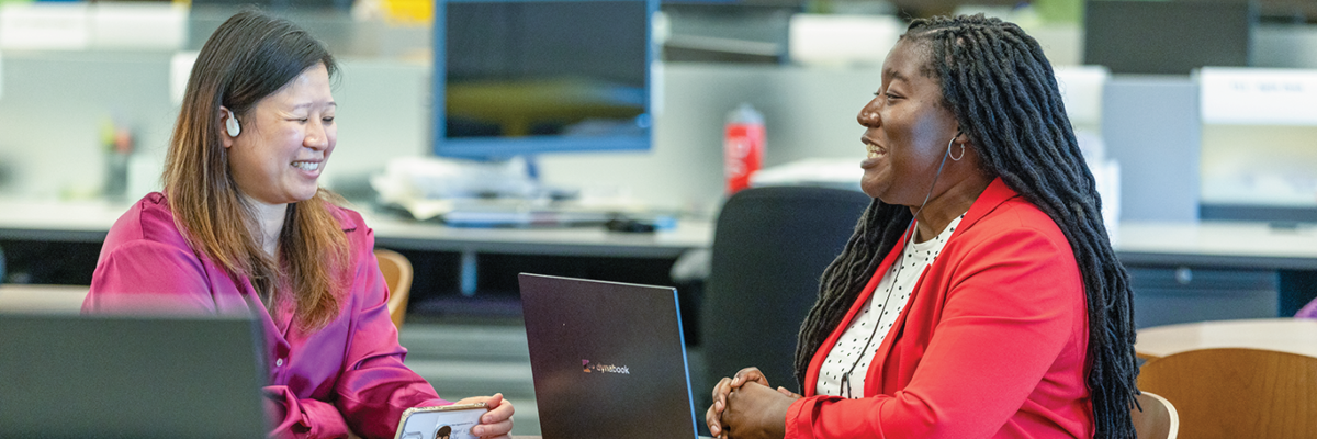 Two women sit at a table in an office environment, engaged in a conversation. The woman on the left is wearing a purple blouse. She is holding a smartphone in her hand and wears earbud headphones. The woman on the right has long braided hair and is wearing a red blazer. Both have laptops on the table in front of them. In the background are computers and different workstations.