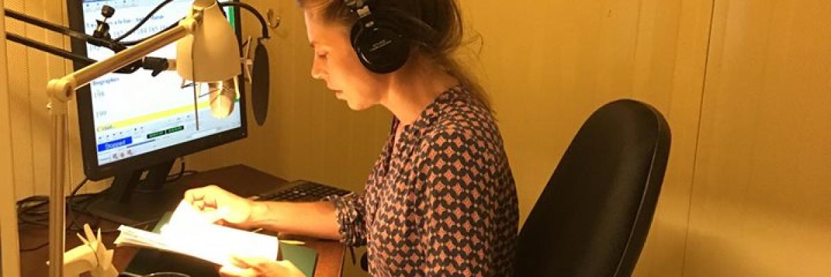 Noémie Godin-Vignault, lectrice bénévole, enregistrant un livre audio.  Elle est assise devant un micro, livre à la main et casque d'écoute sur les oreille dans une cabine d'enregistrement. 