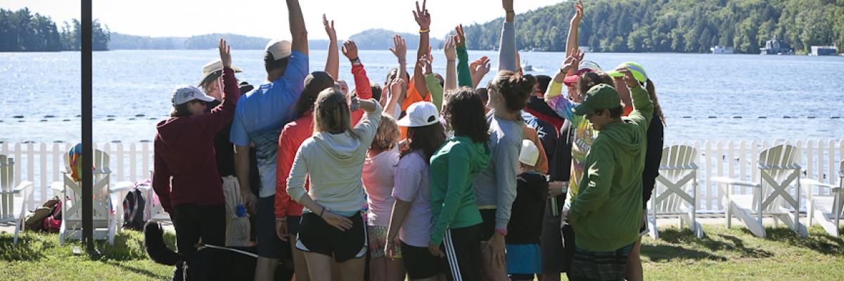 Groupe de jeunes participants du camp, devant un lac, levant les bras en l’air fièrement.