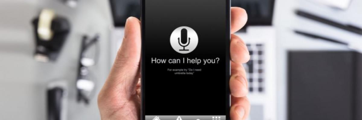 A hand holds an iPhone that’s open to a voice assistant app. It says “How can I help you?”