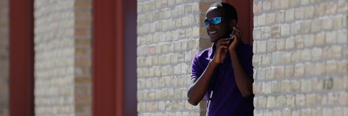 Un homme tient un iPhone contre son oreille. Il porte une chemise violette, des lunettes de soleil et se tient debout contre un mur de briques.