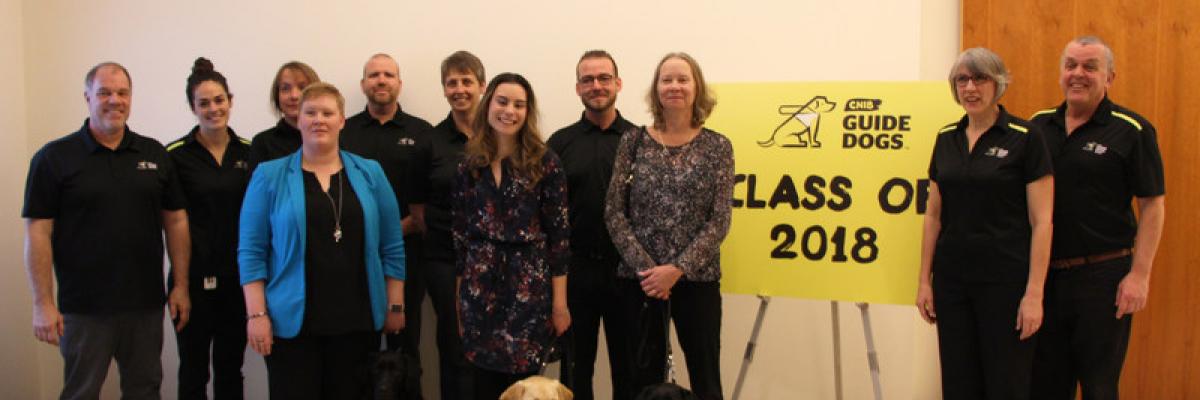 CNIB Guide Dogs graduating class with training staff. A yellow sign in the background reads "Class of 2018"