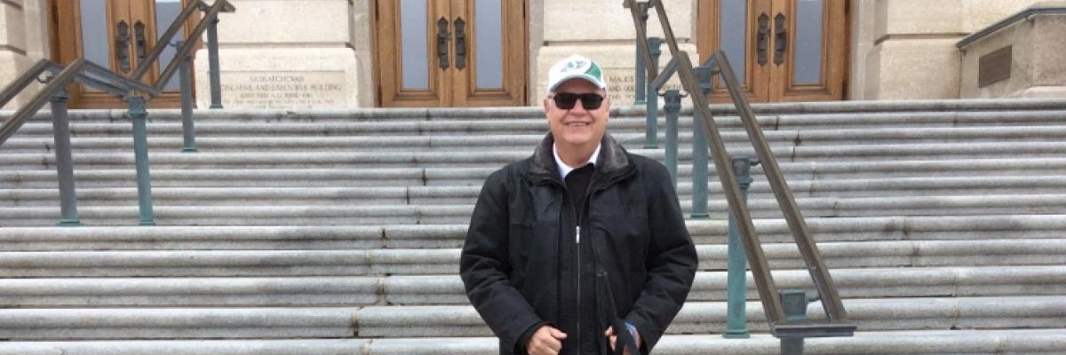 Bob Huber stands outside of the Saskatchewan legislative building with his two dogs, smiling with his sunglasses on and green Roughrider ball cap.