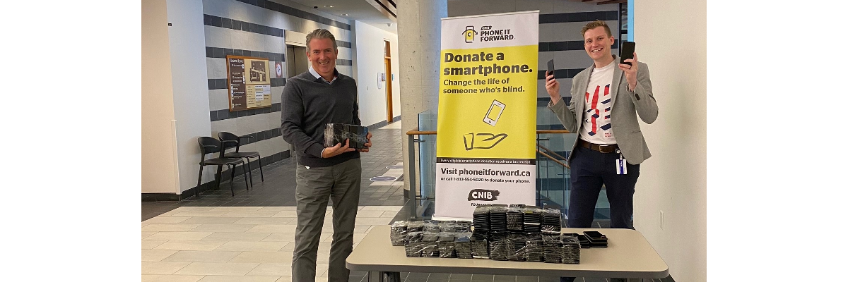 A smiling John M. Rafferty & Thomas Simpson stand at a table and hold multiple donated smartphones from the Bank of Canada. The table is also stacked with donated smartphones. Behind them stands a Phone it Forward banner display.