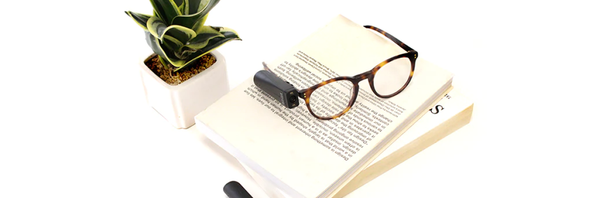 Orcam sur des lunettes déposées sur un livre. Une plante est à gauche du livre.