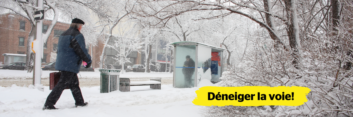 Une rue résidentielle, les trottoirs et les bâtiments environnants sont recouverts de neige. Un homme marche sur un trottoir enneigé. À sa gauche, un abribus et un banc sont également recouverts de neige. Une personne se tient dans l'abribus. Dans le coin inférieur droit de l’image, une bannière jaune affichant le texte « Déneiger la voie! » est superposée.