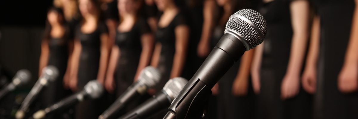 Photo of a number of microphones lined up with a group of women in black dresses in the background.