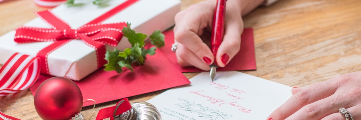 A woman is writing a message inside a card. There are gifts and ornaments and ribbons on the table.