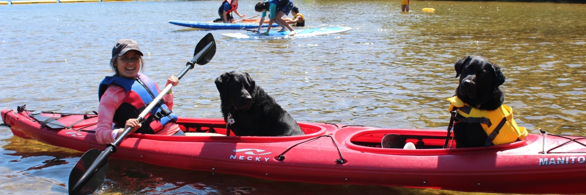 A woman is sitting in a kayak holding a paddle. There are two black guide dogs with her in the boat.