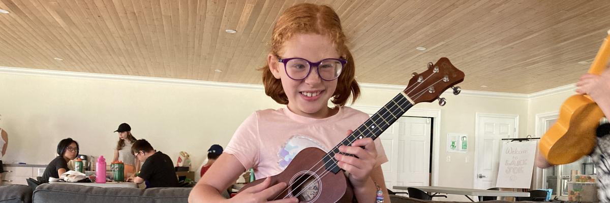 A young girl with red hair and glasses is standing while playing a ukelele in the Rec Hall. She has a big smile on her face.