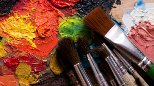 A colourful hand-painted mural with various paintbrushes lying on top of the mural. The abstract mural has thick, textured paint in vibrant colours like red, yellow and green.